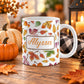 11 oz personalized Changing Leaves Fall Mug with colorful autumn leaves and a custom name in orange script lettering, on a wooden table with fall decor around it including pumpkins, autumn leaves, a candle lantern, and a cozy checkered blanket.