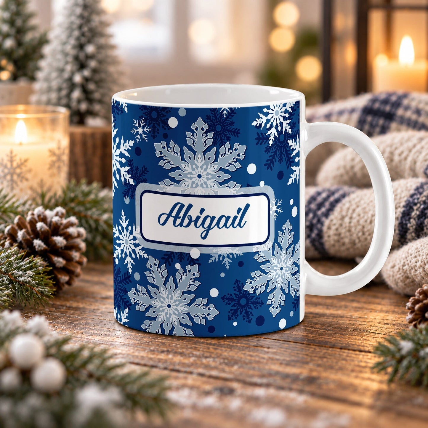 11 oz personalized blue snowflake mug with white and blue winter pattern and custom blue script name in a white panel design on both sides, shown on a wooden table with winter decor around it.