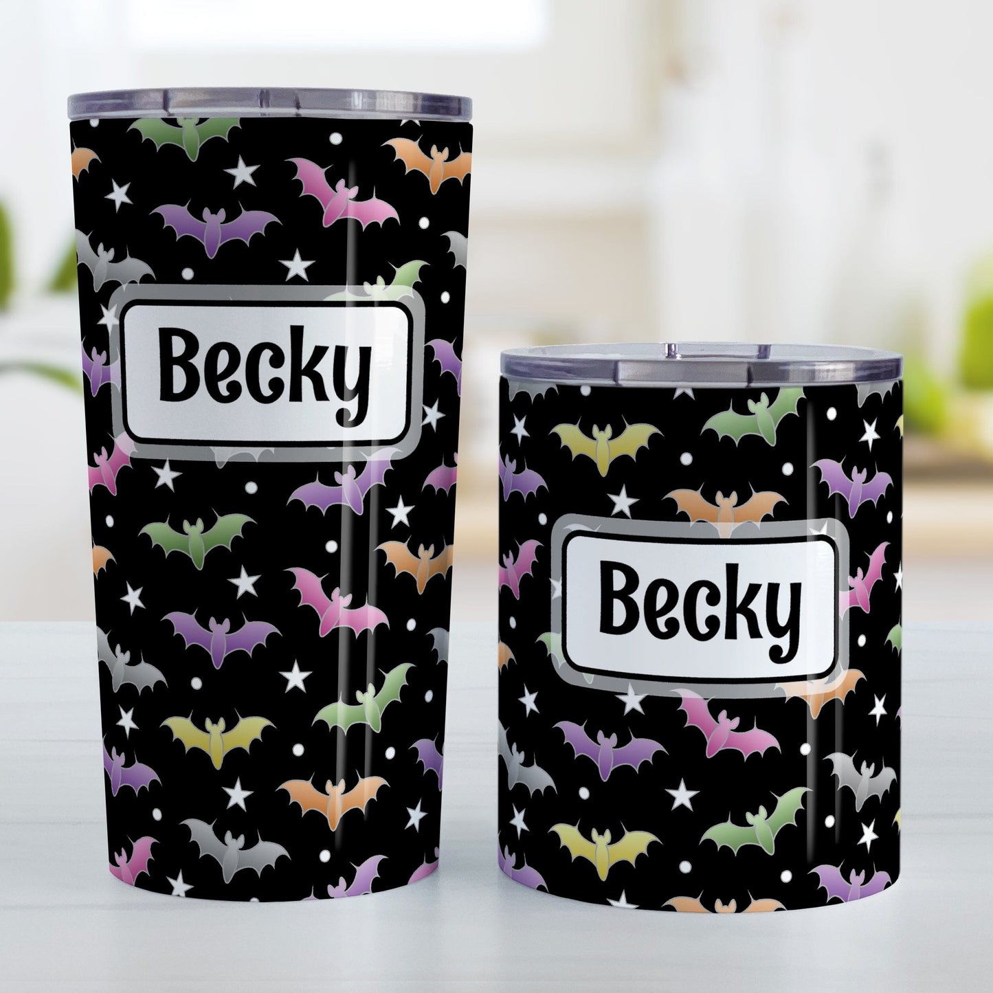 Personalized Colorful Bats Tumbler Cup (20oz or 10oz) at Amy's Coffee Mugs. A tumbler cup designed with different colored bats and white stars in an assorted pattern over a black background color that wraps around the cup. Your personalized name is custom-printed in black within a white rectangular frame design over the colorful bats background. Photo shows both sized cups on a table next to each other.