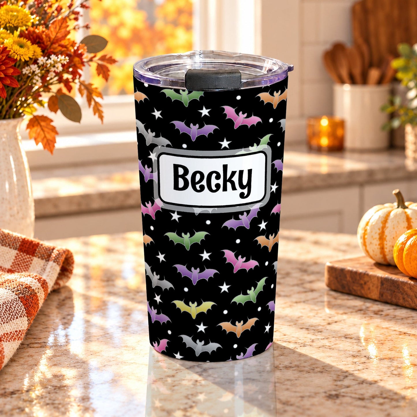 20 oz personalized insulated stainless steel tumbler with colorful bats, white stars and custom black name inside a white frame on a black background, on a granite countertop in a kitchen with fall decor including autumn plants, pumpkins, and an orange plaid tea towel.