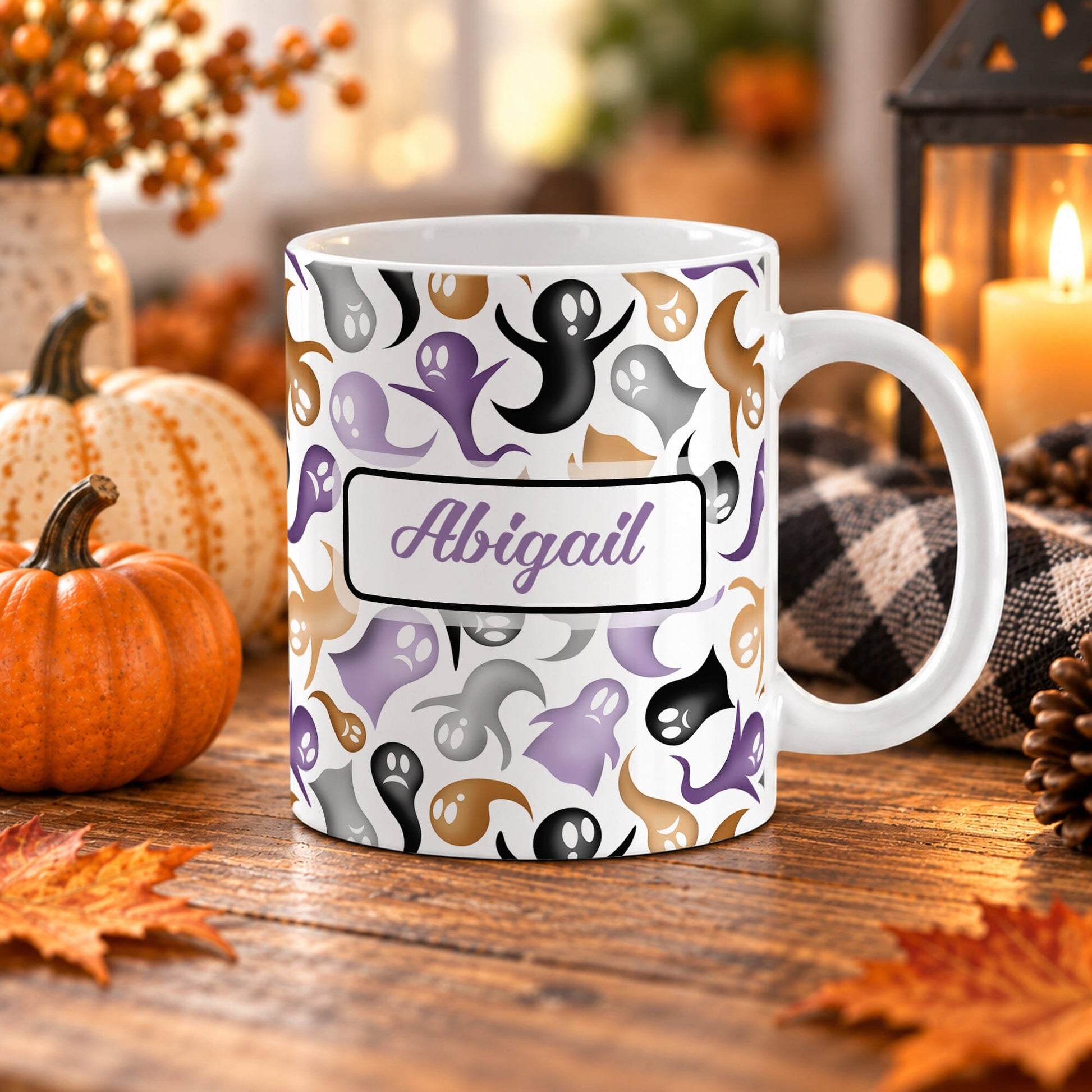 White 11 oz personalized Halloween mug with purple, orange, black, and gray ghosts, plus a custom purple script name in a white bordered panel, on a wooden table with fall decor around it including pumpkins, autumn leaves, a candle lantern, and a cozy checkered blanket.
