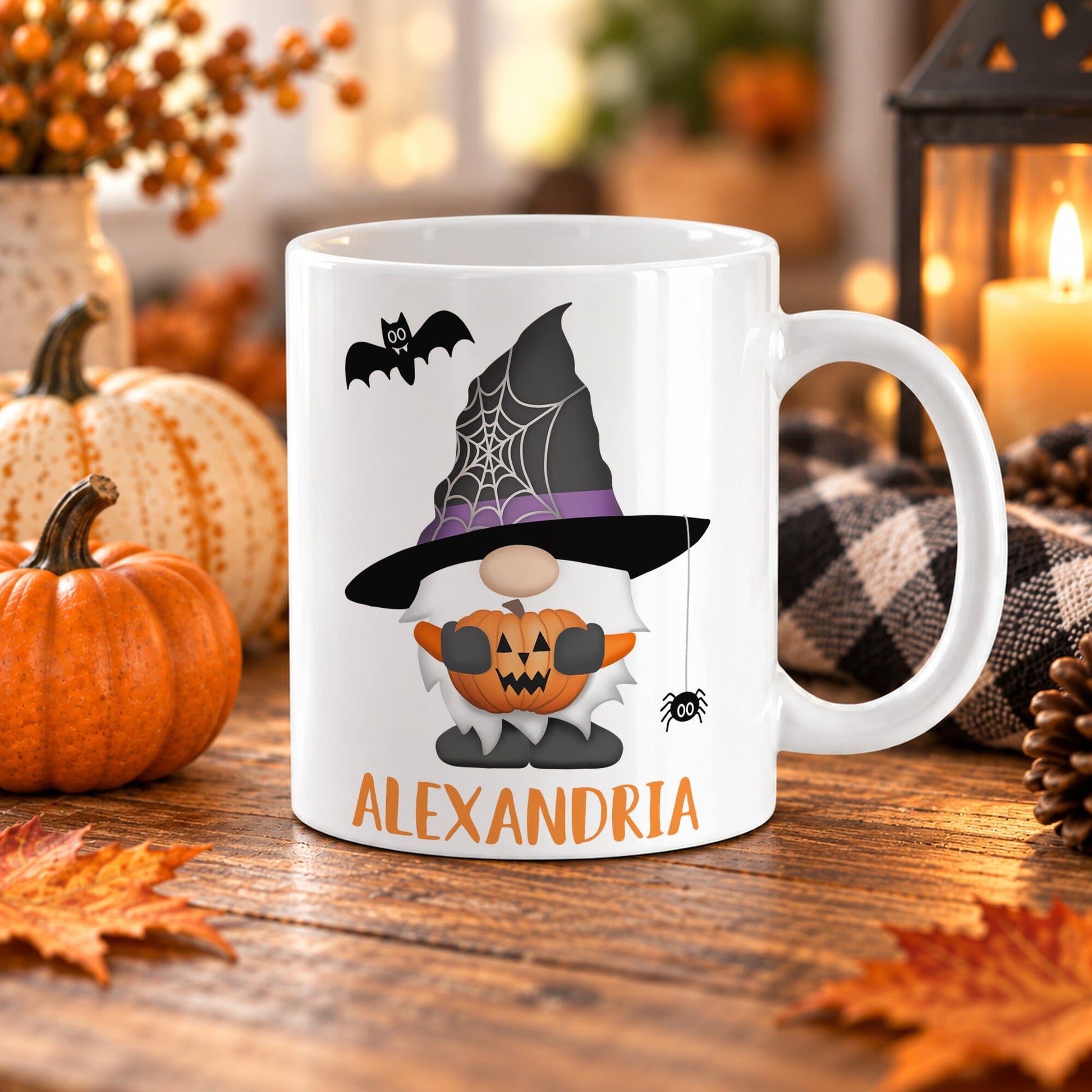 11 oz white ceramic coffee mug with a gnome wearing a witch hat with a dangling spider and a web, holding a carved pumpkin, with a bat overhead, and a personalized name in orange below. The mug is on a wooden table with fall decor around it.