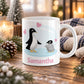 11 oz personalized penguin mug with adult penguin, fluffy baby chick, pink hearts, soft blue background, and custom name in pink, on a wooden table with winter decor around it including pine cones, pine boughs, candles, and a cozy plaid blanket.