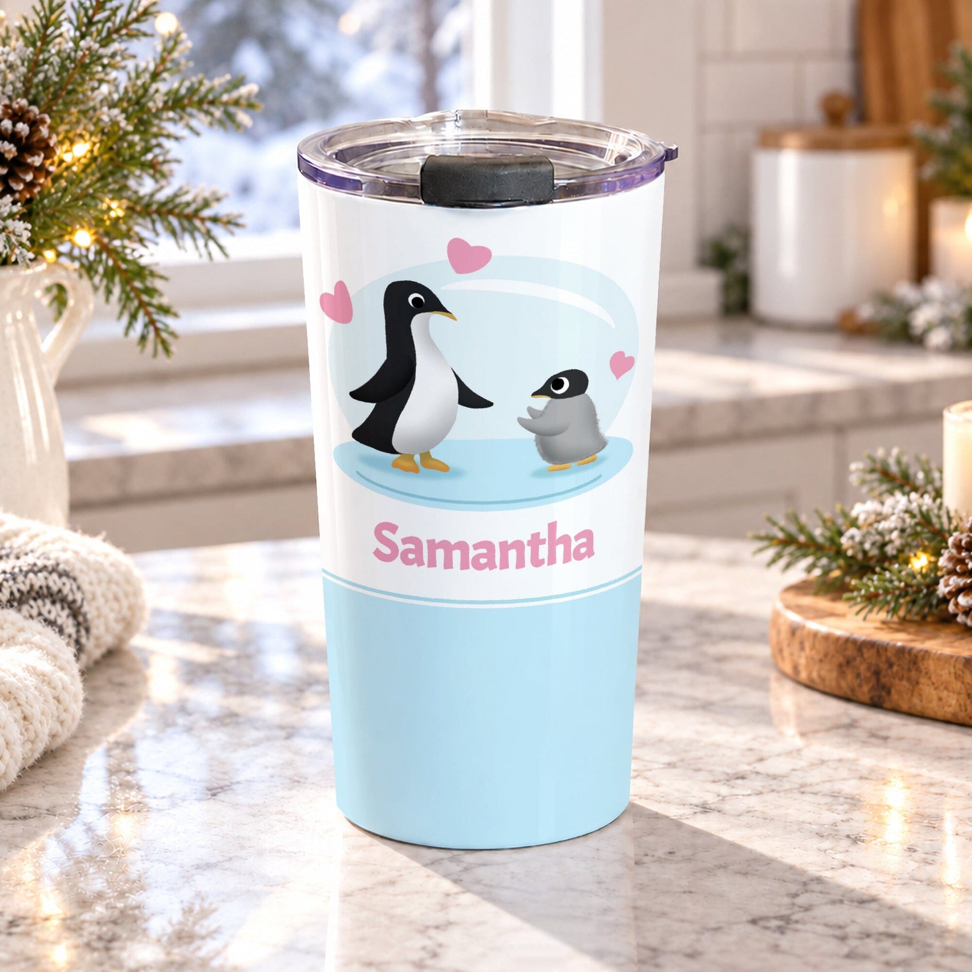 20 oz personalized penguin tumbler cup with parent and baby chick, pink hearts, custom name in pink, and soft blue design, on a granite countertop in a kitchen with light rustic winter decor including pine cones, pine boughs, and a cozy blanket.