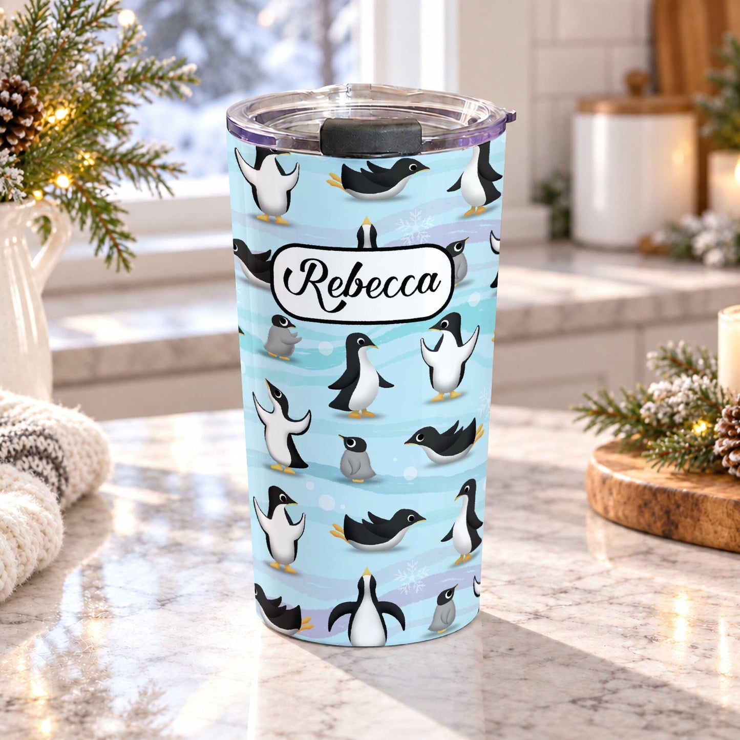 20 oz personalized penguin tumbler cup with adult and baby penguins, blue winter pattern, snowflakes, and custom name in black script, on a granite countertop in a light rustic kitchen with pine cones, pine boughs, lights, and a cozy blanket.