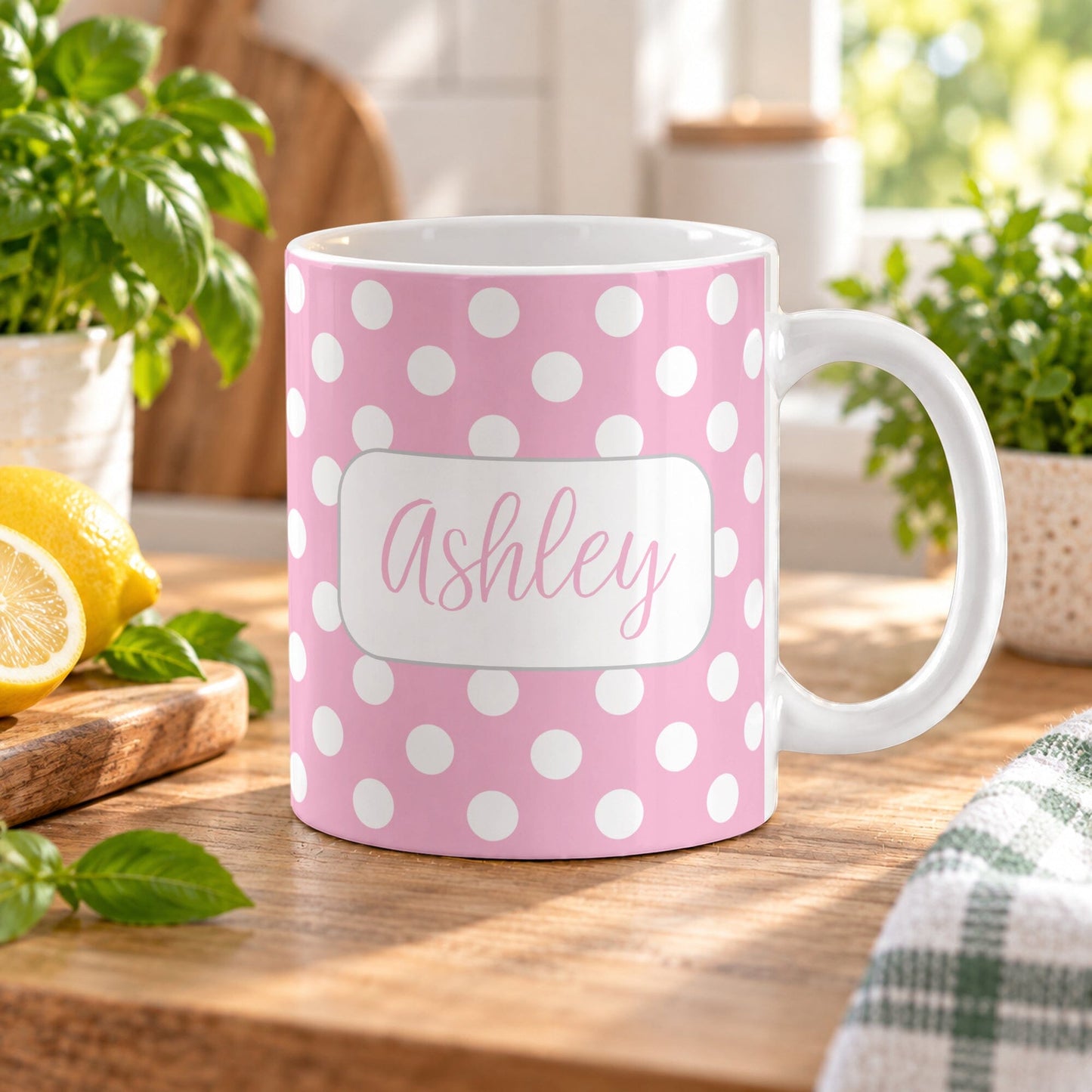 11 oz personalized pink polka dot mug with white dots and custom name on both sides, displayed on a wooden table in a bright kitchen with green plants, a tea towel, and lemons on a cutting board.