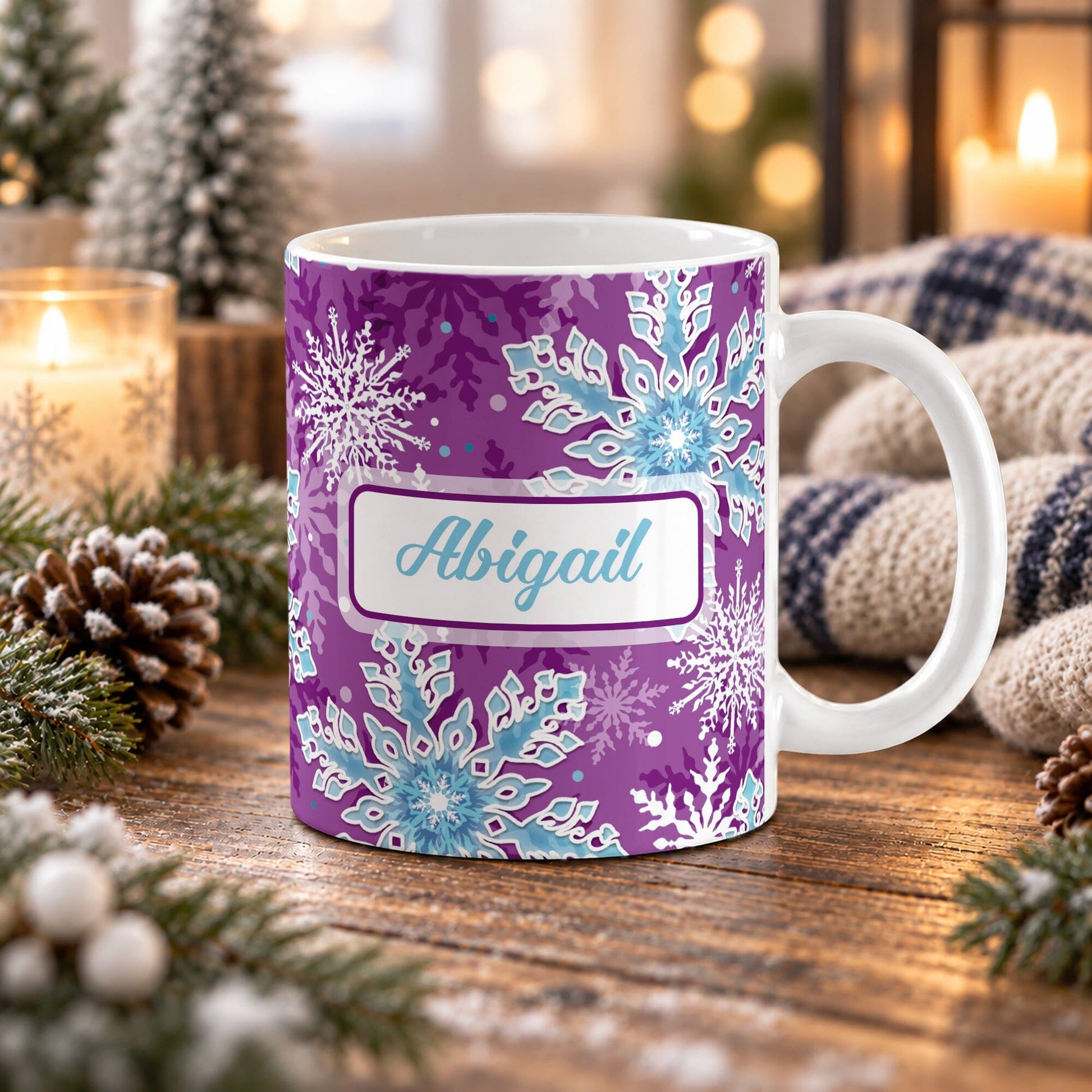 11 oz personalized purple snowflake mug with blue and white snowflakes, aqua blue custom name, and purple winter background, on a wooden table with winter decor around it including pine cones, pine boughs, candles, and a cozy plaid blanket.