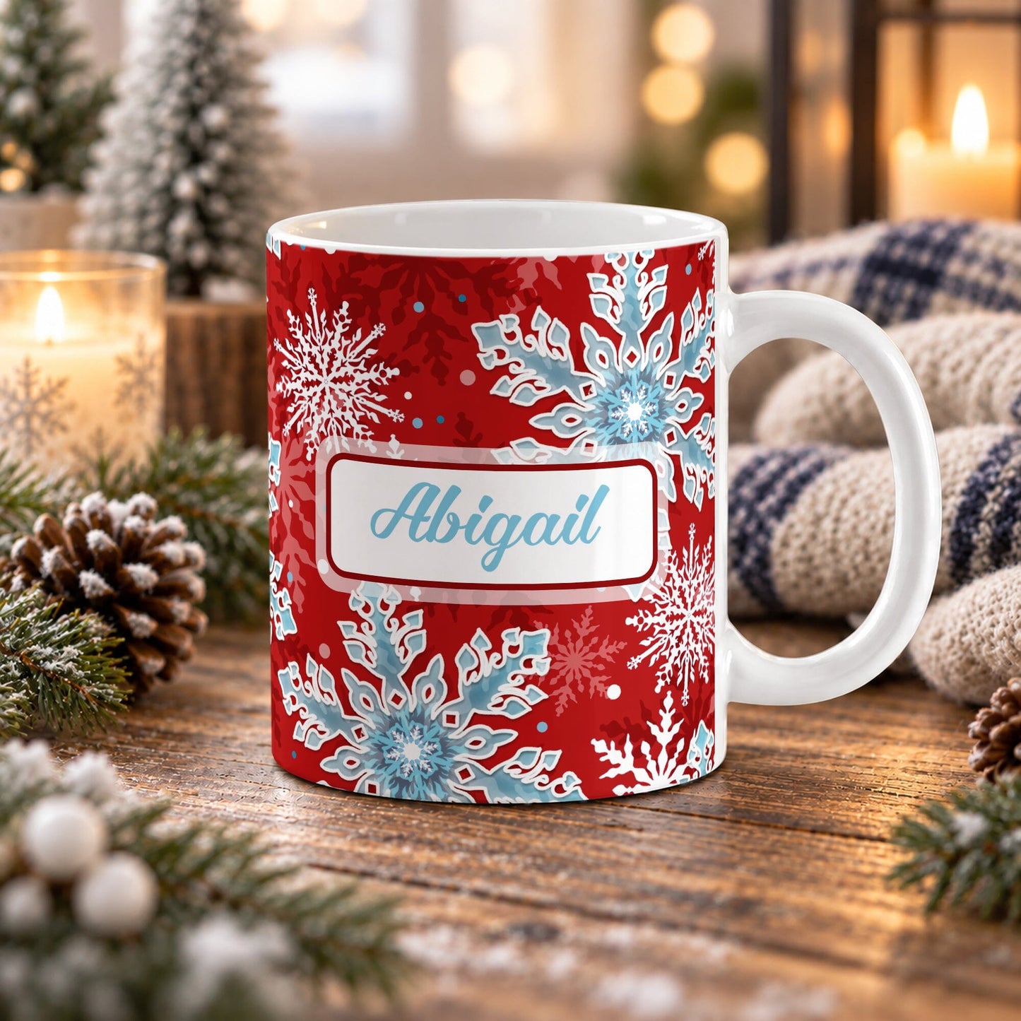 11 oz personalized red and blue snowflake mug with blue custom name, layered winter snowflakes, and red background, on a wooden table with winter decor around it including pine cones, pine boughs, candles, and a cozy plaid blanket.