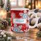 11 oz personalized red and blue snowflake mug with blue custom name, layered winter snowflakes, and red background, on a wooden table with winter decor around it including pine cones, pine boughs, candles, and a cozy plaid blanket.