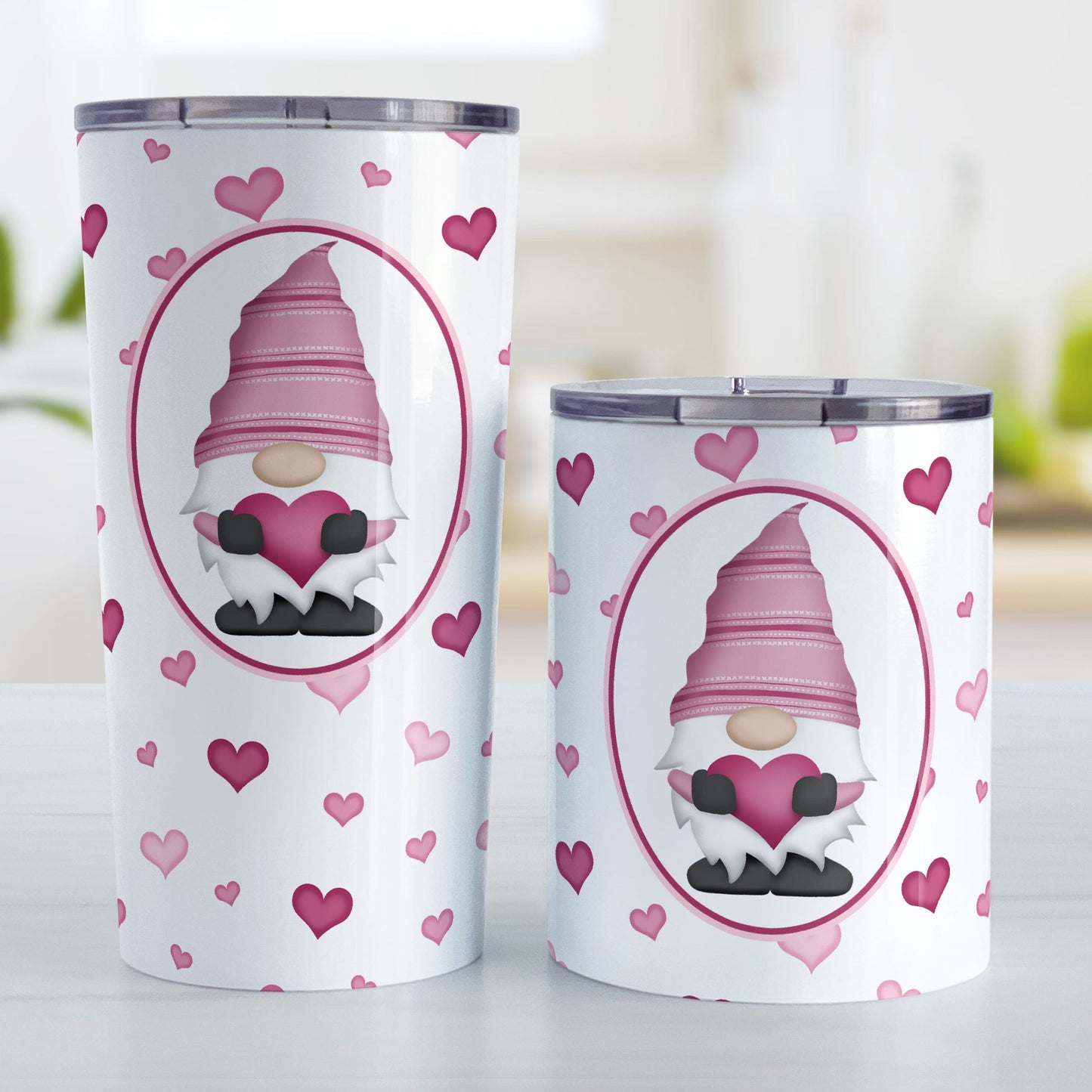 Pink Gnome Dainty Hearts Tumbler Cup (20oz or 10oz) at Amy's Coffee Mugs. Stainless steel tumbler cups designed with an adorable pink gnome holding a heart in a white oval over a pattern of cute and dainty hearts in different shades of pink that wrap around the cups. Photo shows both sized cups on a table next to each other. 