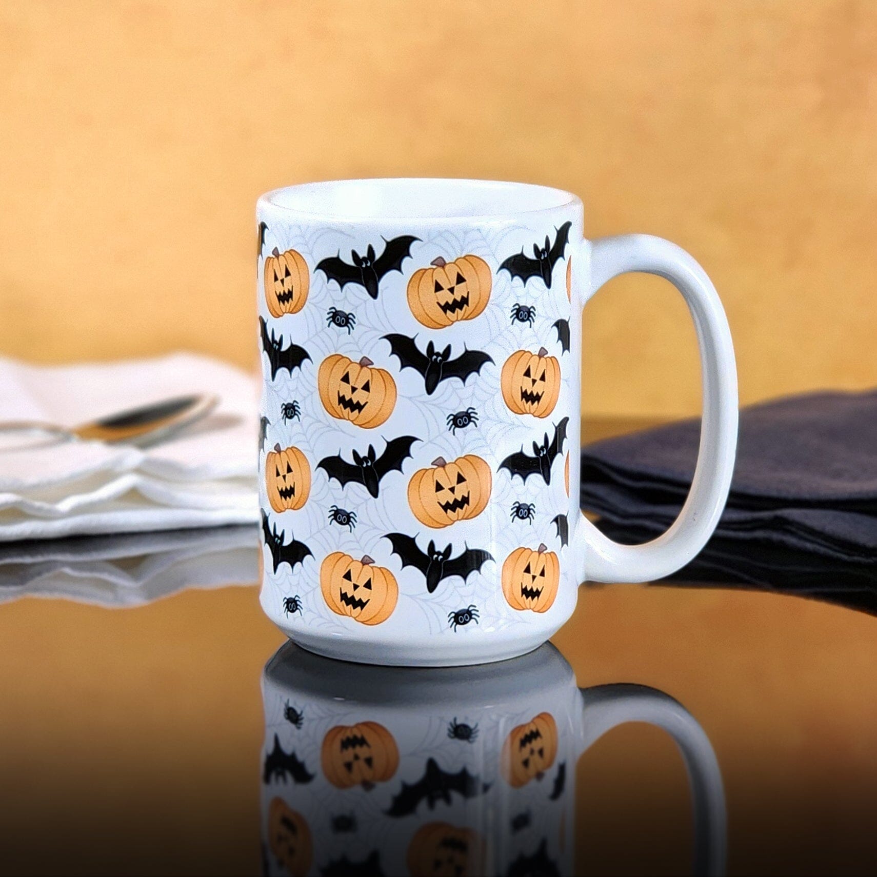 Pumpkins Bats and Spiders Halloween Mug (15oz) at Amy's Coffee Mugs. Photo shows the mug on a glossy black table with an orange background.