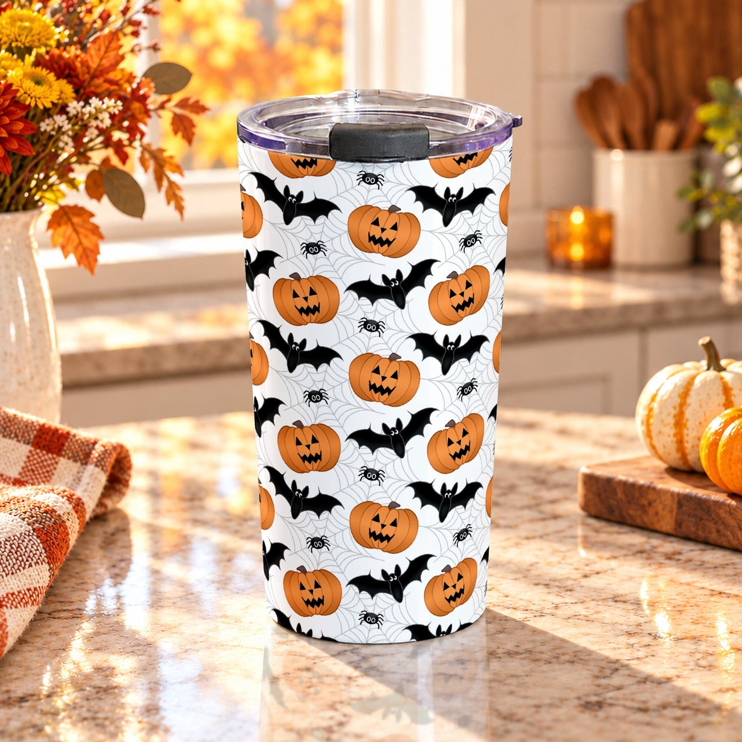 20 oz insulated stainless steel Halloween tumbler cup with orange jack-o'-lanterns, black bats, spiders, and light gray spider webs on white, on a granite countertop in a kitchen with fall decor including autumn plants, pumpkins, and an orange plaid tea towel.