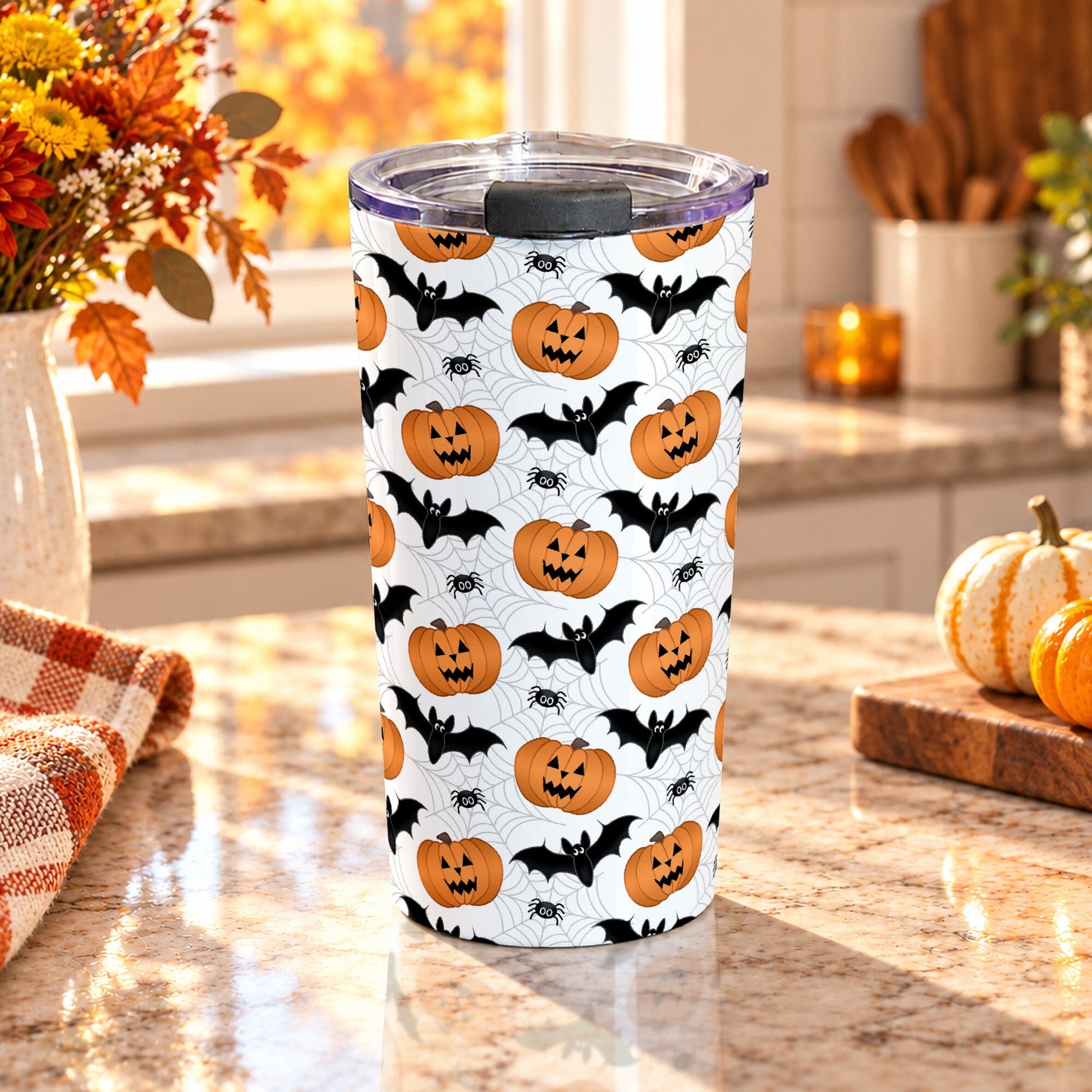 20 oz insulated stainless steel Halloween tumbler cup with orange jack-o'-lanterns, black bats, spiders, and light gray spider webs on white, on a granite countertop in a kitchen with fall decor including autumn plants, pumpkins, and an orange plaid tea towel.