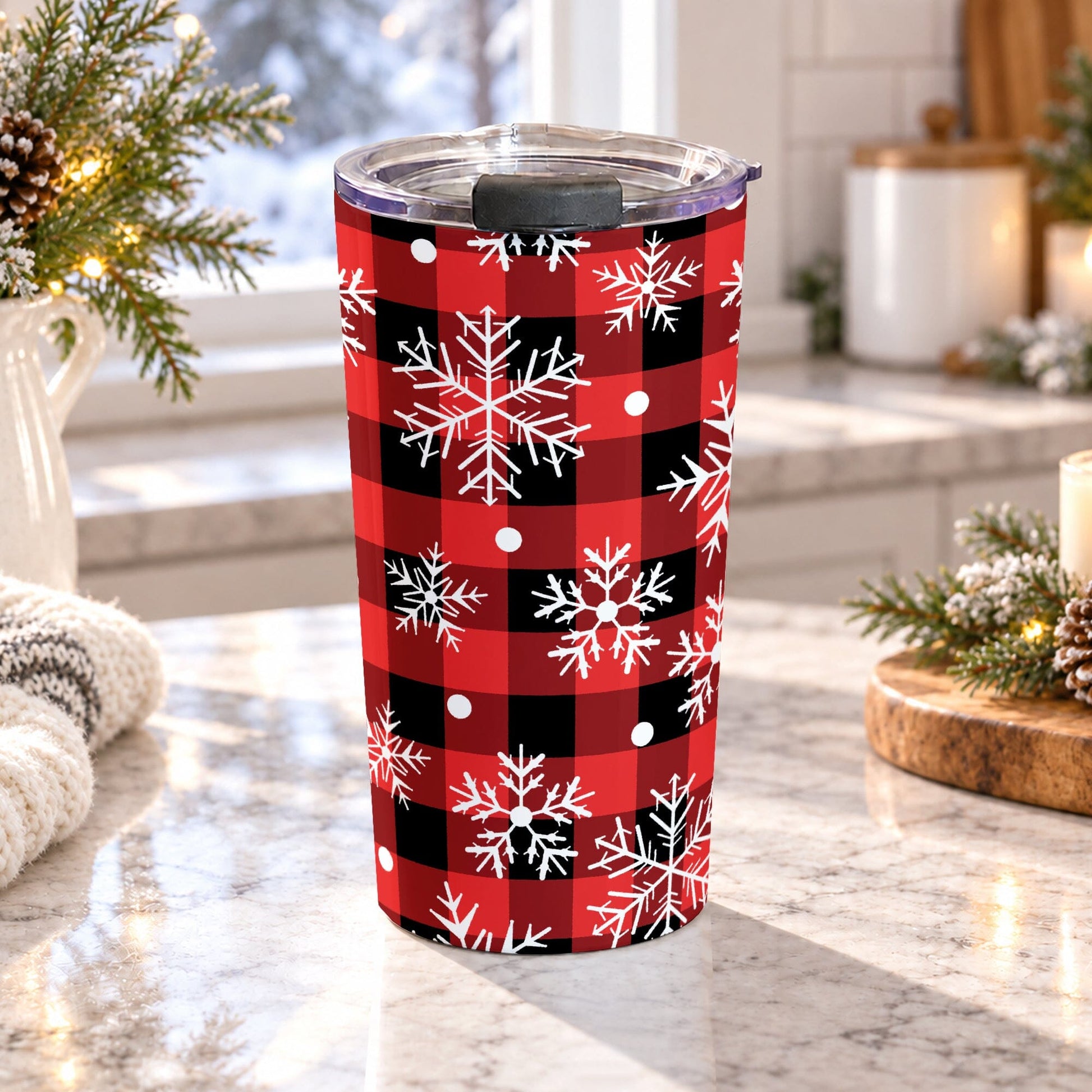20 oz red and black buffalo plaid snowflake tumbler cup with white snowflakes and snowy dot accents, on a granite countertop in a bright kitchen with light rustic winter decor including pine cones, pone boughs, and a cozy blanket.