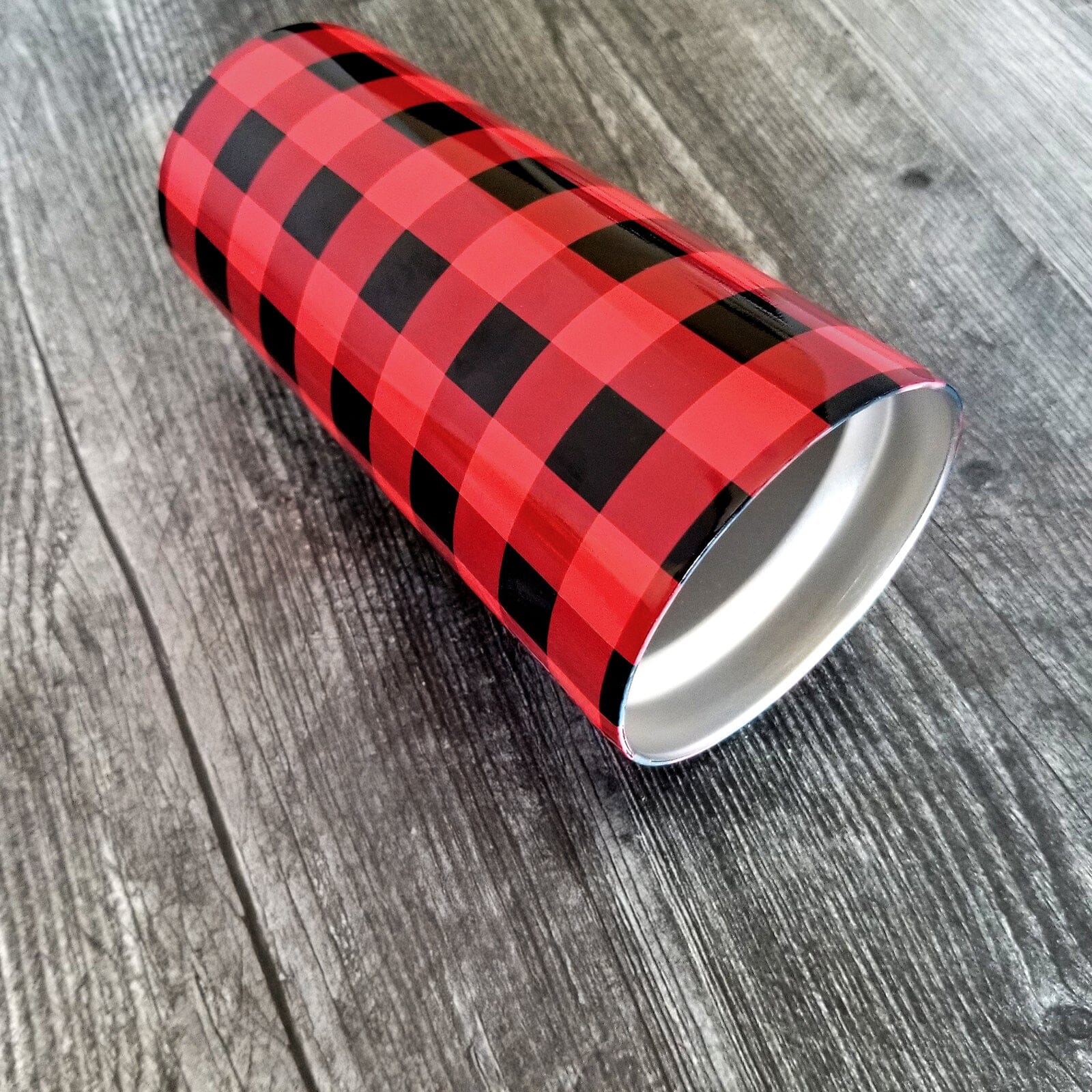 A 20oz tumbler cup designed with a red and black buffalo plaid (buffalo check) pattern that wraps around the cup. Photo shows the cup laying on its side on a weathered wood surface.