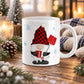 An 11 oz white ceramic mug featuring a festive holiday winter gnome design with a red and black buffalo plaid hat, white beard, black shoes, and a red snow shovel, set on a wooden table surrounded by candles, pinecones, and a cozy knit blanket.