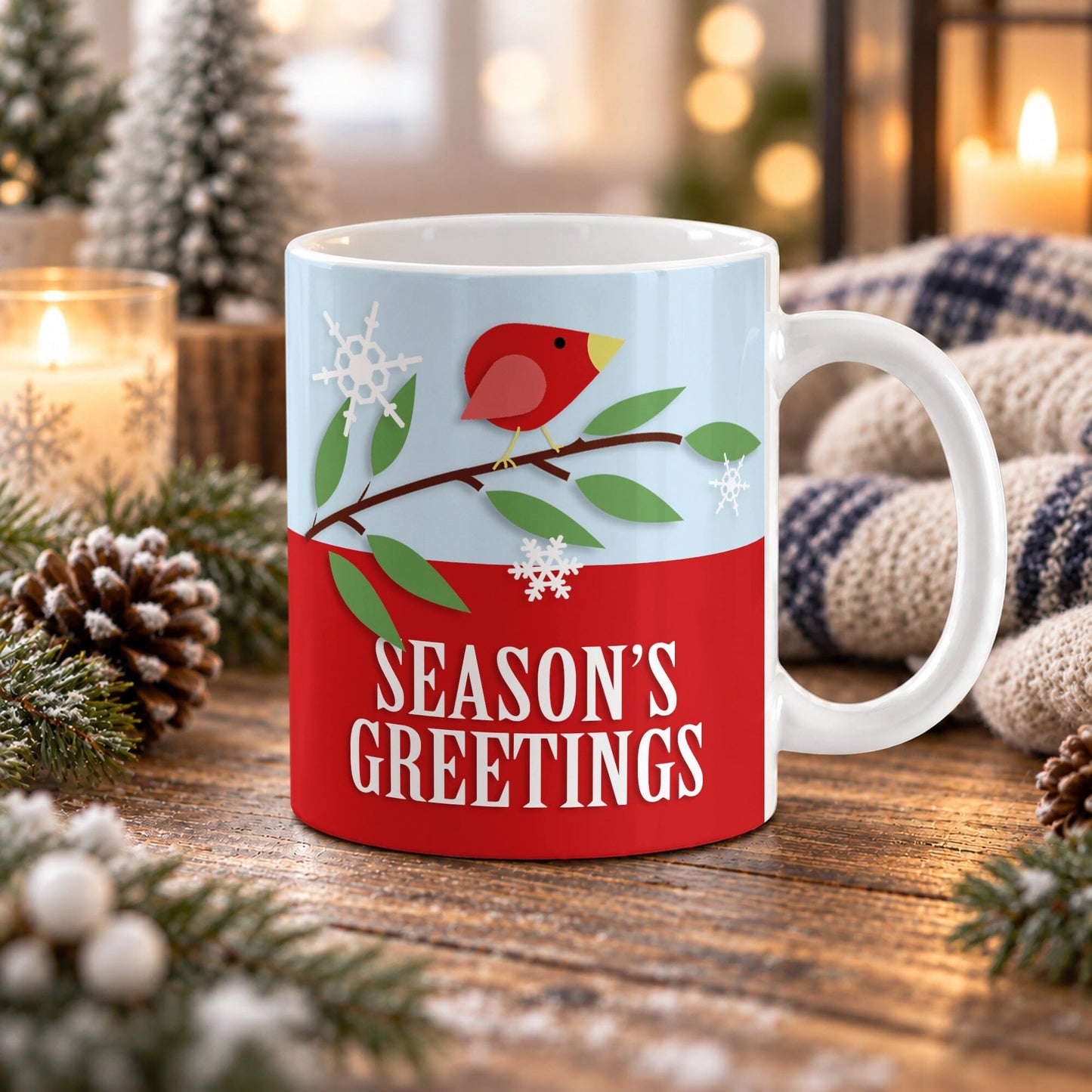 11 oz Christmas mug with red bird on a leafy branch, snowflakes, and Season’s Greetings text on a red and light blue design, on a wooden table with winter decor around it including pine cones, pin boughs, candles, and a cozy plaid blanket.