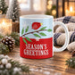 11 oz Christmas mug with red bird on a leafy branch, snowflakes, and Season’s Greetings text on a red and light blue design, on a wooden table with winter decor around it including pine cones, pin boughs, candles, and a cozy plaid blanket.