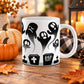 White 11 oz ceramic Halloween mug with black rising ghosts, gravestones, crosses, coffin-shaped markers, and RIP cemetery details, on a wooden table with fall decor including pumpkins, autumn leaves, a candle lantern, and a cozy checkered blanket.