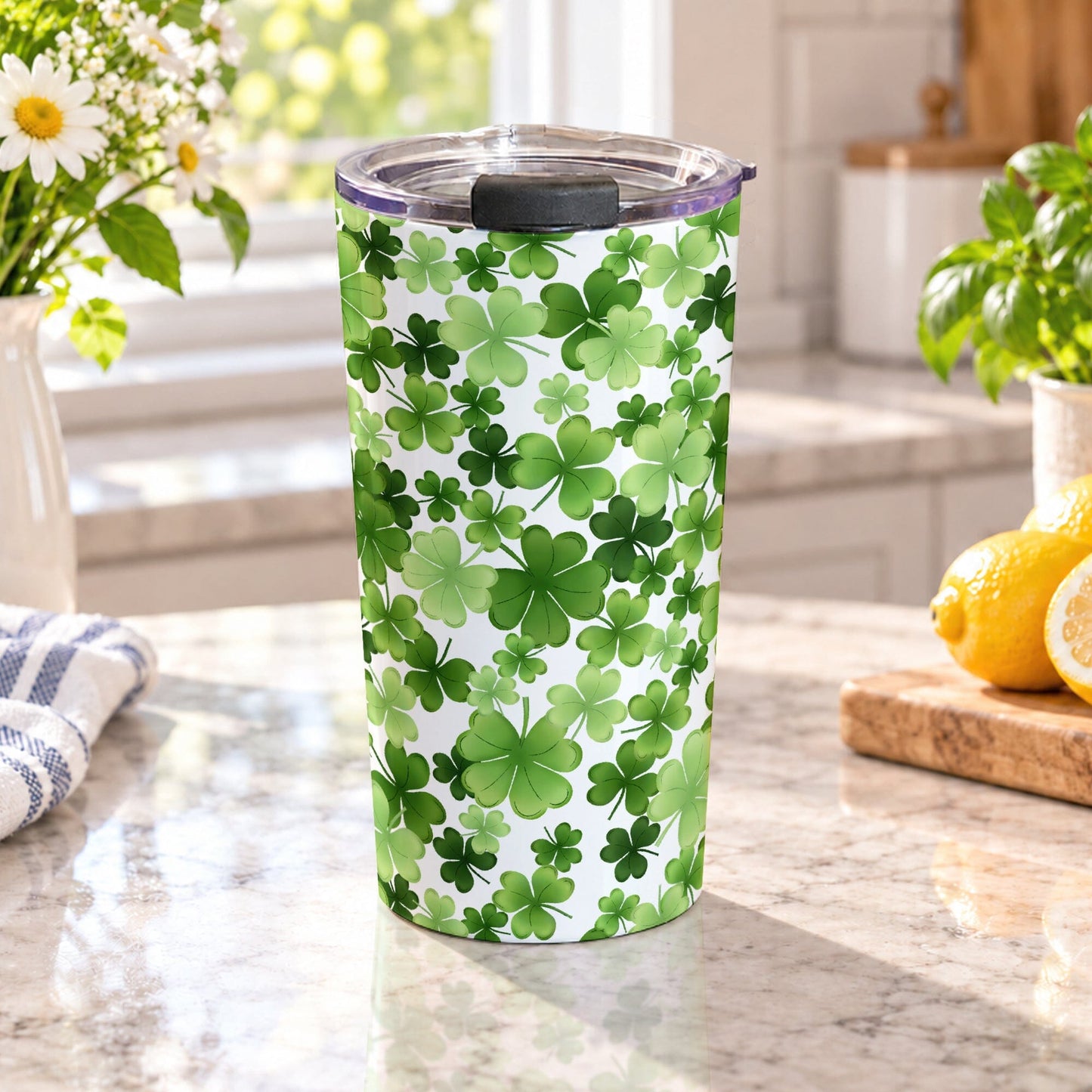 20 oz insulated stainless steel tumbler cup covered with green shamrocks and four-leaf clovers in multiple shades on a white background, on a granite countertop in a bright kitchen with green plants, daisies, a plaid tea towel, and lemons on a wood cutting board.