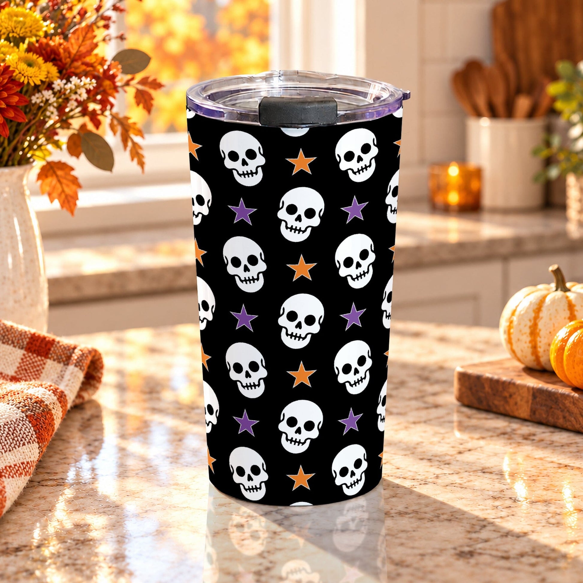 20 oz insulated stainless steel Halloween tumbler cup with repeating white skulls and orange and purple stars on a black background, on a granite countertop in a kitchen with fall decor including autumn plants, pumpkins, and an orange plaid tea towel.