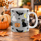 White 11 oz ceramic Halloween mug with black cat, white ghost, jack-o'-lantern, black bat, crescent moon, spiders and spider webs on gray, shown on a wooden table with fall decor.