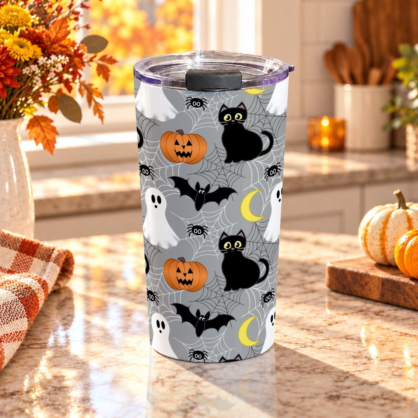 20 oz insulated stainless steel Halloween tumbler cup with black cat, white ghost, jack-o'-lantern, bat, crescent moon, spiders, and spider webs on gray, on a granite countertop in a kitchen with fall decor including autumn plants, pumpkins, and an orange plaid tea towel.
