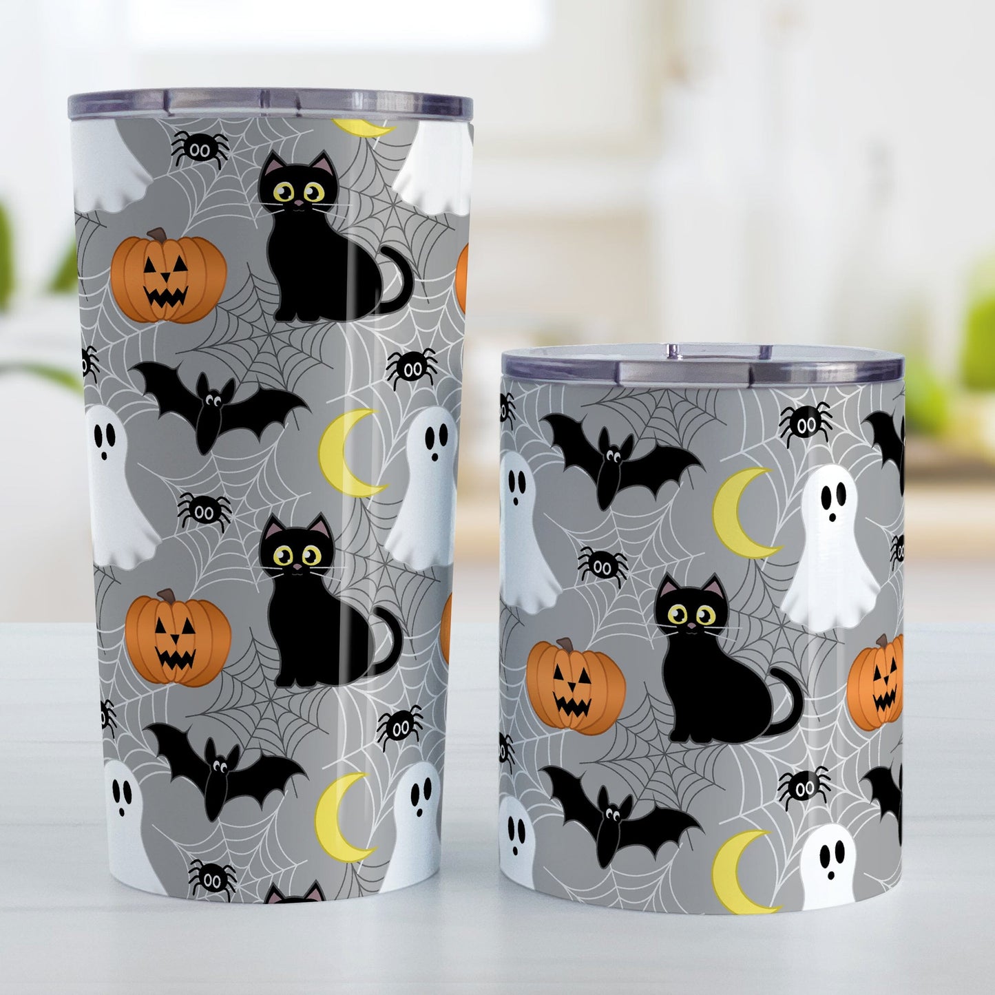 Spooky Cute Halloween Tumbler Cup (20oz or 10oz) at Amy's Coffee Mugs. A tumbler cup designed with a cute black cat, ghost, jack o'lantern, crescent moon, bat, spider, and spider webs over a gray background color in a pattern that wraps around the cup. Photo shows both sized cups on a table next to each other.