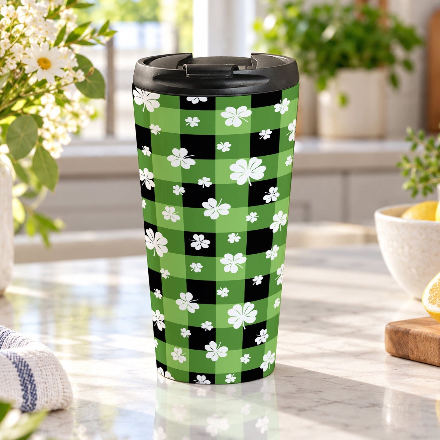 15 oz green and black buffalo plaid travel mug with white shamrocks and four-leaf clovers, displayed in a bright kitchen on a granite countertop with green plants, daisies, a teal towel, and lemons in a bowl and on a cutting board.
