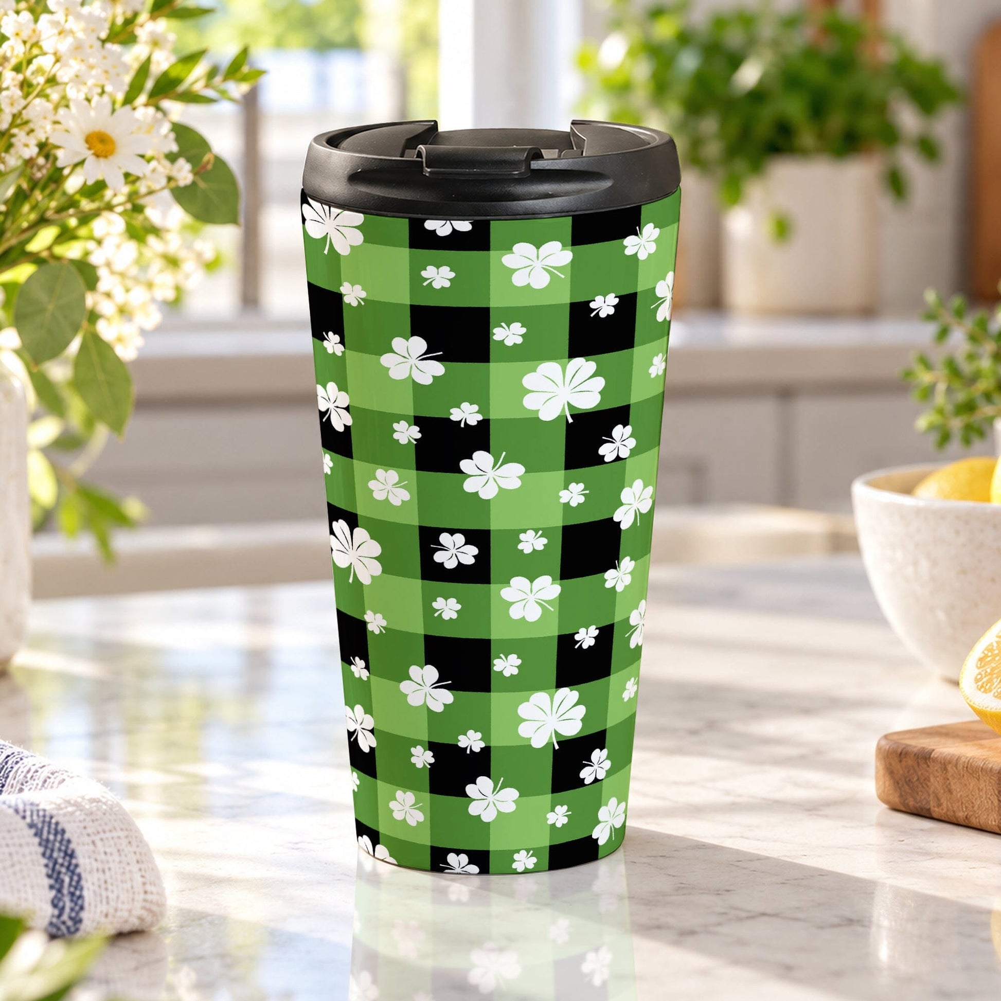 15 oz green and black buffalo plaid travel mug with white shamrocks and four-leaf clovers, displayed in a bright kitchen on a granite countertop with green plants, daisies, a teal towel, and lemons in a bowl and on a cutting board.