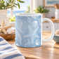 11 oz blue beach mug with white line-art seashells, scallop shells, spiral shells, conch shells, and starfish in a coastal pattern, set on a wooden table in a bright, coastal-themed kitchen.