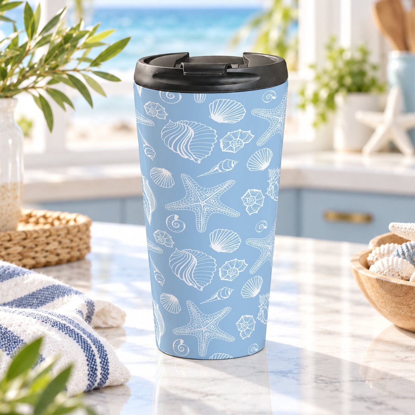 15 oz blue beach travel mug with white line-art seashells, starfish, and coastal shell illustrations, displayed on a granite countertop in a beach house kitchen with coastal decor.