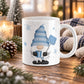 11 oz winter gnome mug with blue snowflake hat, blue coffee mug, light blue snow shovel, and snowflakes on white ceramic, displayed on a wooden table with winter decor around it.