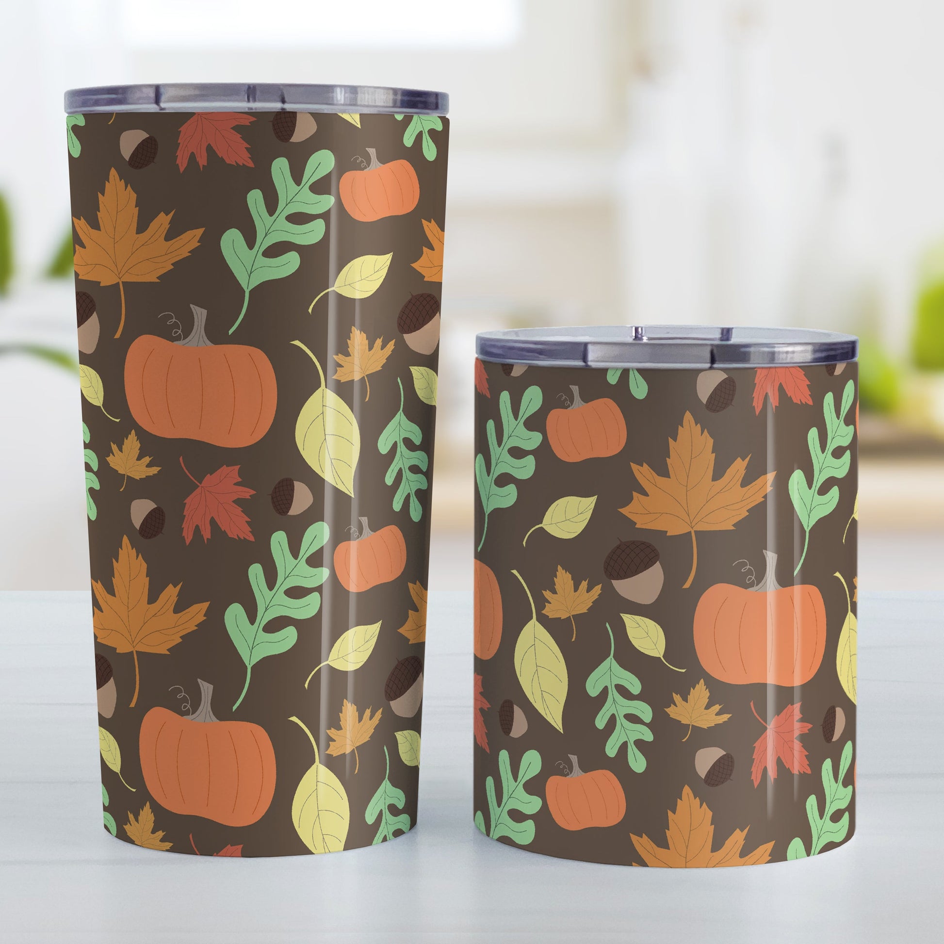 Autumn Pumpkins and Leaves Pattern - Fall Tumbler Cup (20oz and 10oz, stainless steel insulated) at Amy's Coffee Mugs. Tumbler cups designed with a pattern of pumpkins, acorns, and leaves in orange, yellow, and green over a brown background color that wraps around the cups. Photo shows both sized cups on a table next to each other.