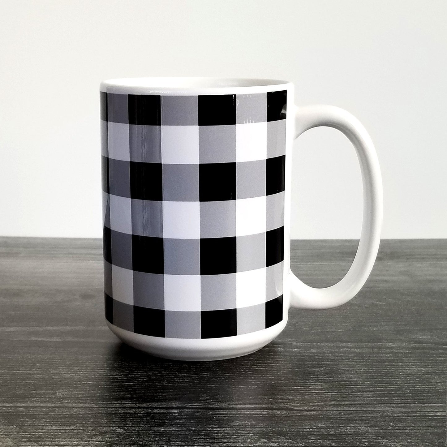 Black and White Buffalo Plaid Mug (15oz, on white and weathered wood background) at Amy's Coffee Mugs. A ceramic coffee mug designed with a black and white buffalo plaid (buffalo check) pattern that wraps around the mug to the handle. Photo shows the mug sitting on a dark wood table.