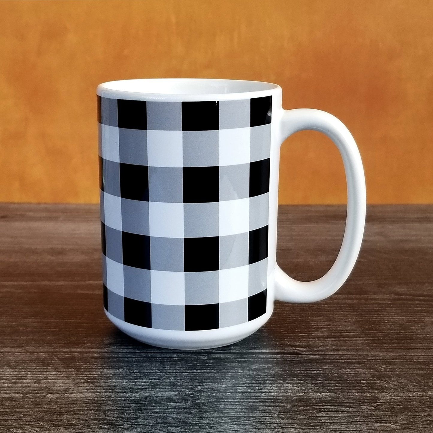 Black and White Buffalo Plaid Mug (15oz, on orange and weathered wood background) at Amy's Coffee Mugs. A ceramic coffee mug designed with a black and white buffalo plaid (buffalo check) pattern that wraps around the mug to the handle. Photo shows the mug sitting on a dark wood table with an orange background.