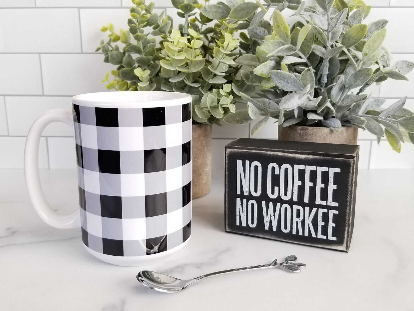 Black and White Buffalo Plaid Mug at Amy's Coffee Mugs. A ceramic coffee mug designed with a black and white buffalo plaid (buffalo check) pattern that wraps around the mug to the handle. Photo shows the mug sitting on a countertop next to a coffee sign, a spoon, and plants. 