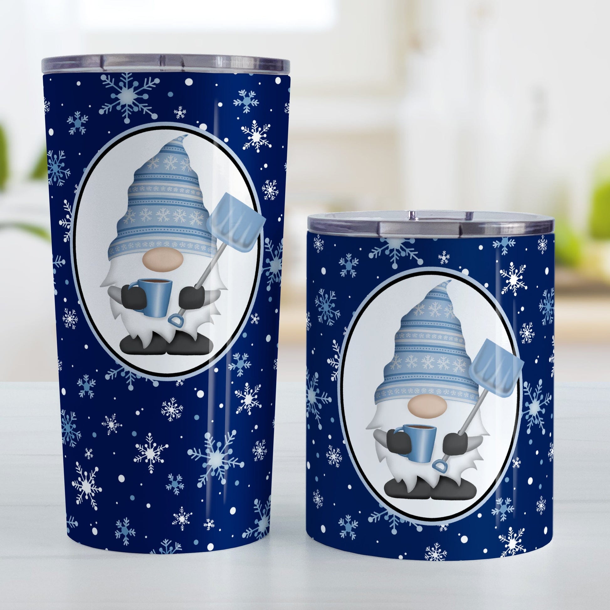 Blue Gnome Snowflakes Tumbler Cup (20oz or 10oz) at Amy's Coffee Mugs. Stainless steel tumbler cups designed with an adorable gnome wearing a festive blue winter hat and holding a hot beverage and a snow shovel in a white oval over a blue night snowflakes background that wraps around the cups. Photo shows both sized cups next to each other on a table.
