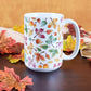 Changing Leaves Fall Mug (15oz) on a cherry wood table with leaves and a beige linen towel in front of an orange wall. 