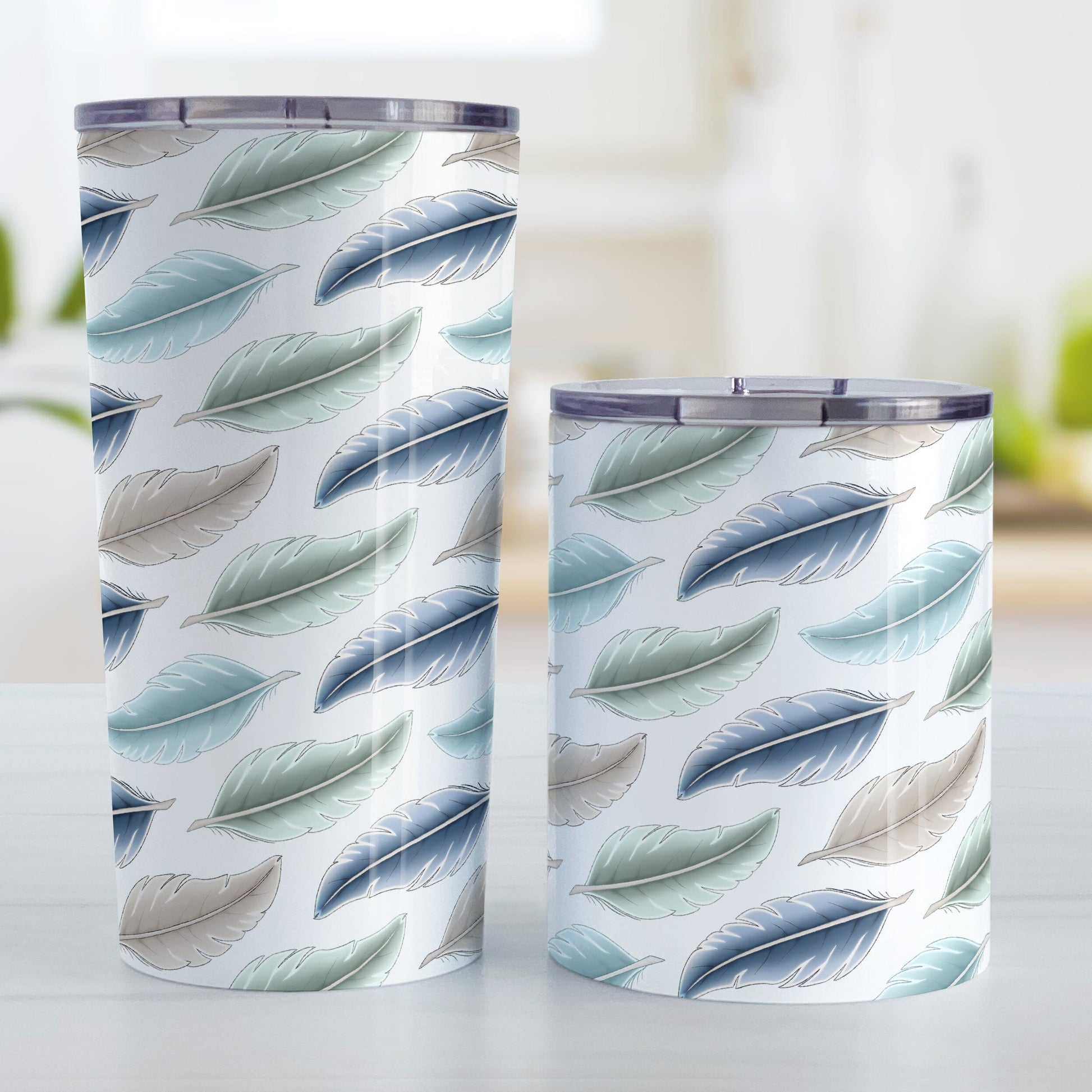 Coastal Feathers Tumbler Cup (20oz or 10oz) at Amy's Coffee Mugs. Stainless steel tumbler cups designed with a pattern of feathers in a coastal color scheme that wraps around the cups. Photo shows both sized cups next to each other on a table.