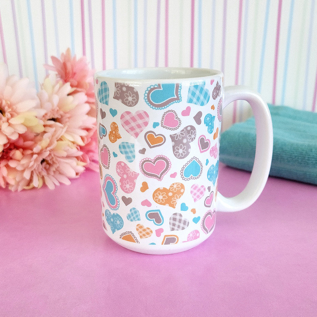 Cutesy Hearts Pattern Mug (15oz) at Amy's Coffee Mugs. This ceramic coffee mug is designed with adorable little hearts in pink, turquoise, orange, and brown. This pattern of cute hearts wraps around the mug to the handle. Photo shows the mug on a pink table with flowers and a towel, in front of a cute striped wall. 
