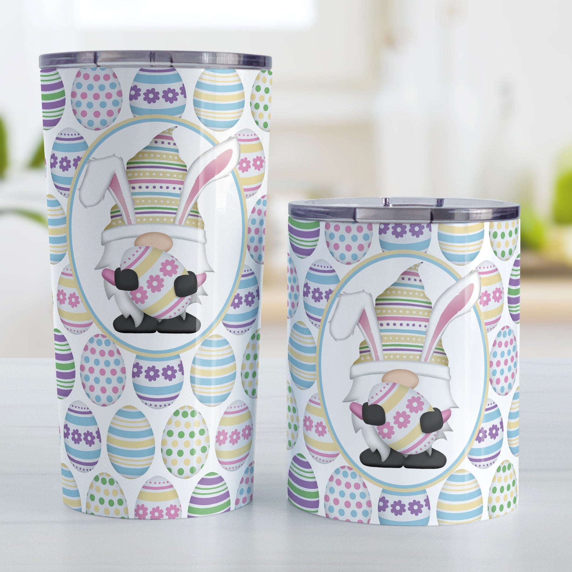 Easter Gnome Eggs Tumbler Cup (20oz or 10oz) at Amy's Coffee Mugs. Stainless steel tumbler cups designed with an illustration of an adorable gnome with bunny ears, holding a large pink and yellow Easter egg, in a white oval over a decorated Easter eggs pattern that wraps around the cups. Photo shows both sized cups next to each other on a table. 