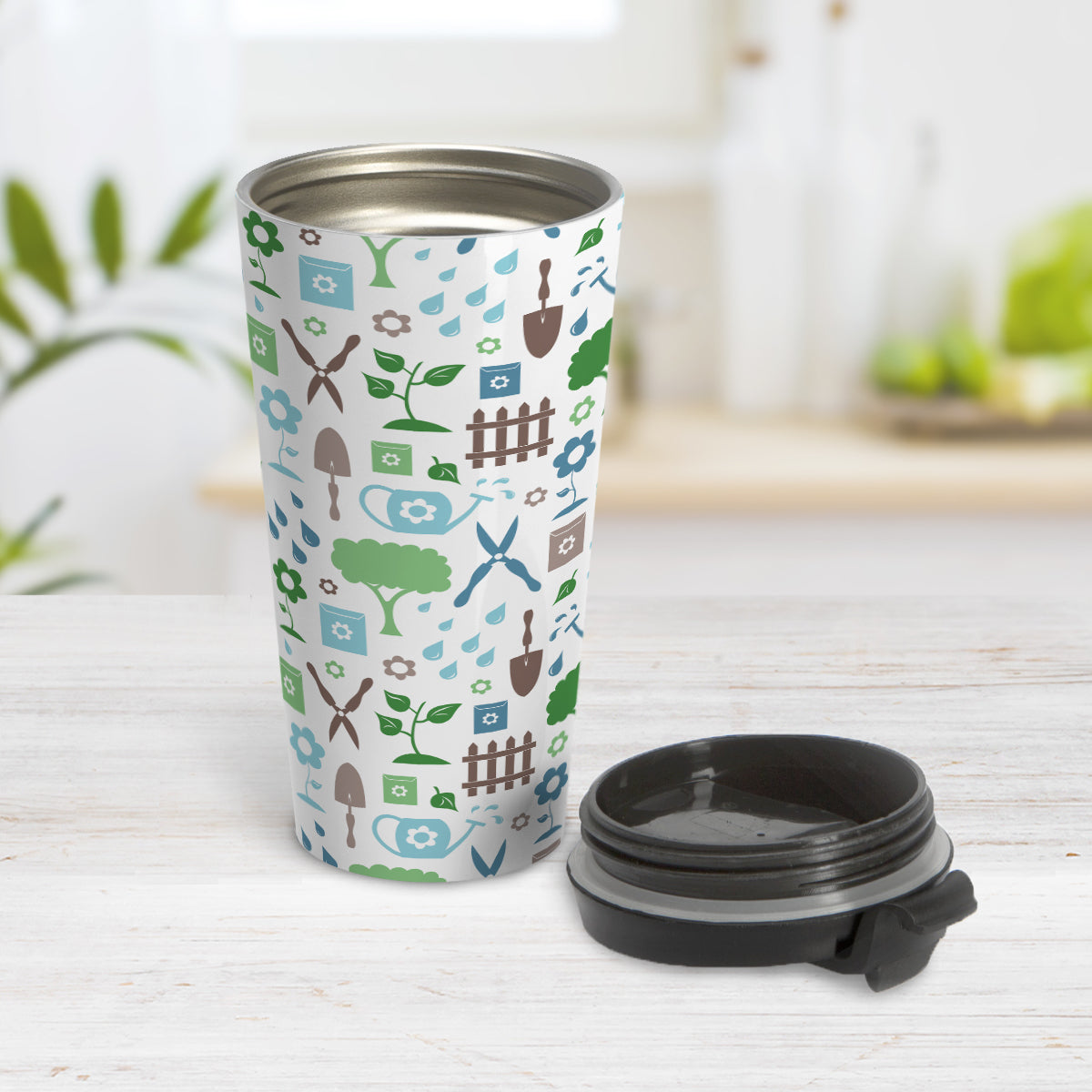 Gardening Pattern Travel Mug (15oz) at Amy's Coffee Mugs. A stainless steel travel mug designed with a gardening pattern with trees, plants, flowers, seed packets, watering cans, fences, and gardening tools in blue, green, and brown. Photo shows the mug open with the lid on the table beside it.