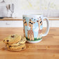 Gathering Giraffes Mug (15oz) at Amy's Coffee Mugs. A ceramic coffee mug designed with five cute illustrated giraffes, with different expressions, around the mug, over a blue sky and green grass background. Photo shows the mug on a kitchen island next to cookies. 