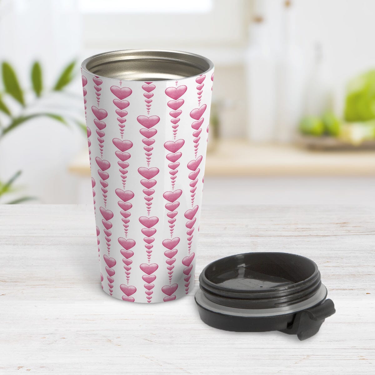 Heart Strings Travel Mug at Amy's Coffee Mugs. A stainless steel insulated travel mug designed with strings of pink hearts descending in size in a pattern that wraps around the mug. Photo shows the travel mug open with the lid on the table beside it.