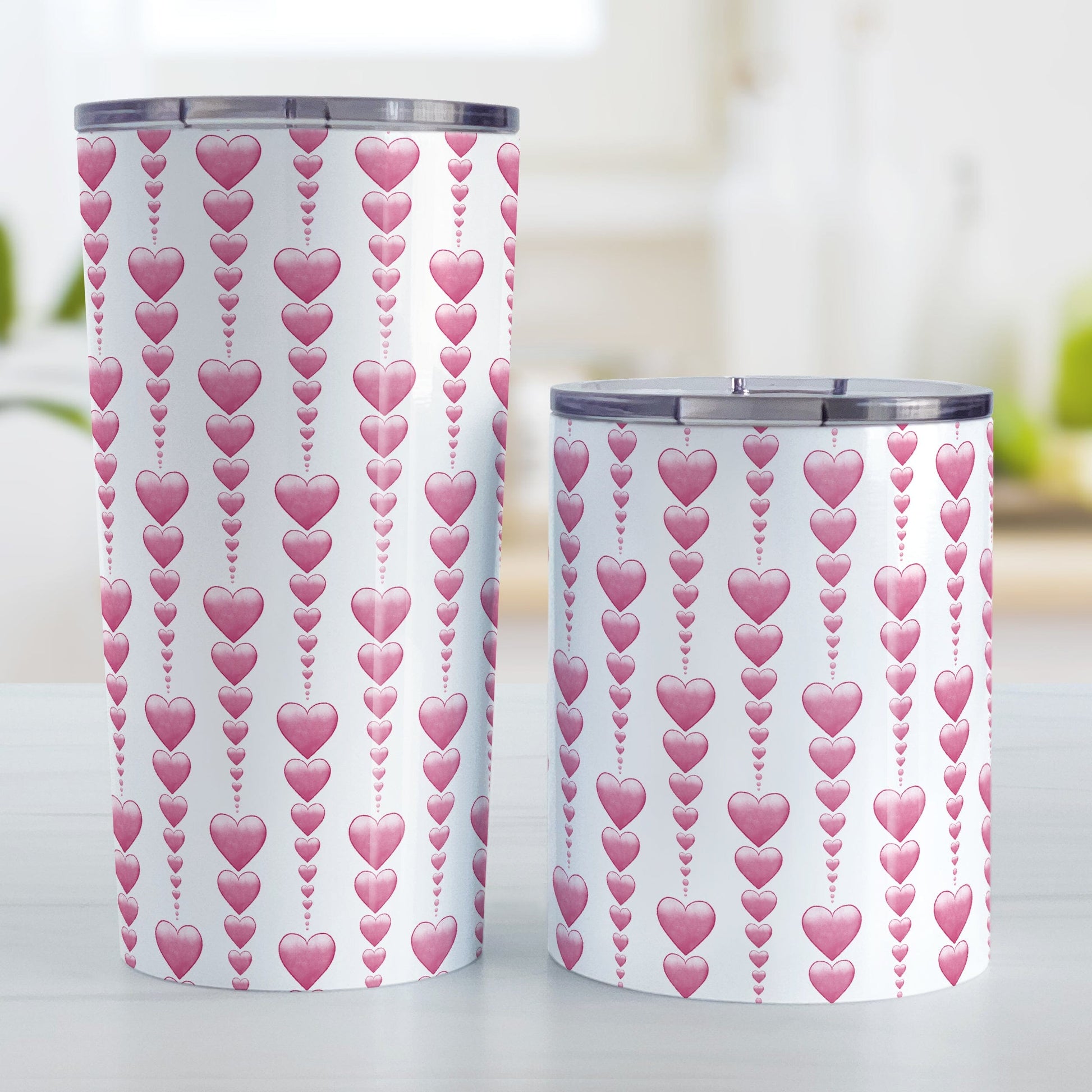 Heart Strings Tumbler Cup (20oz or 10oz) at Amy's Coffee Mugs. Stainless steel insulated tumbler cups designed with strings of pink hearts descending in size in a pattern that wraps around the cups. Photo shows both sized cups next to each other on a table.
