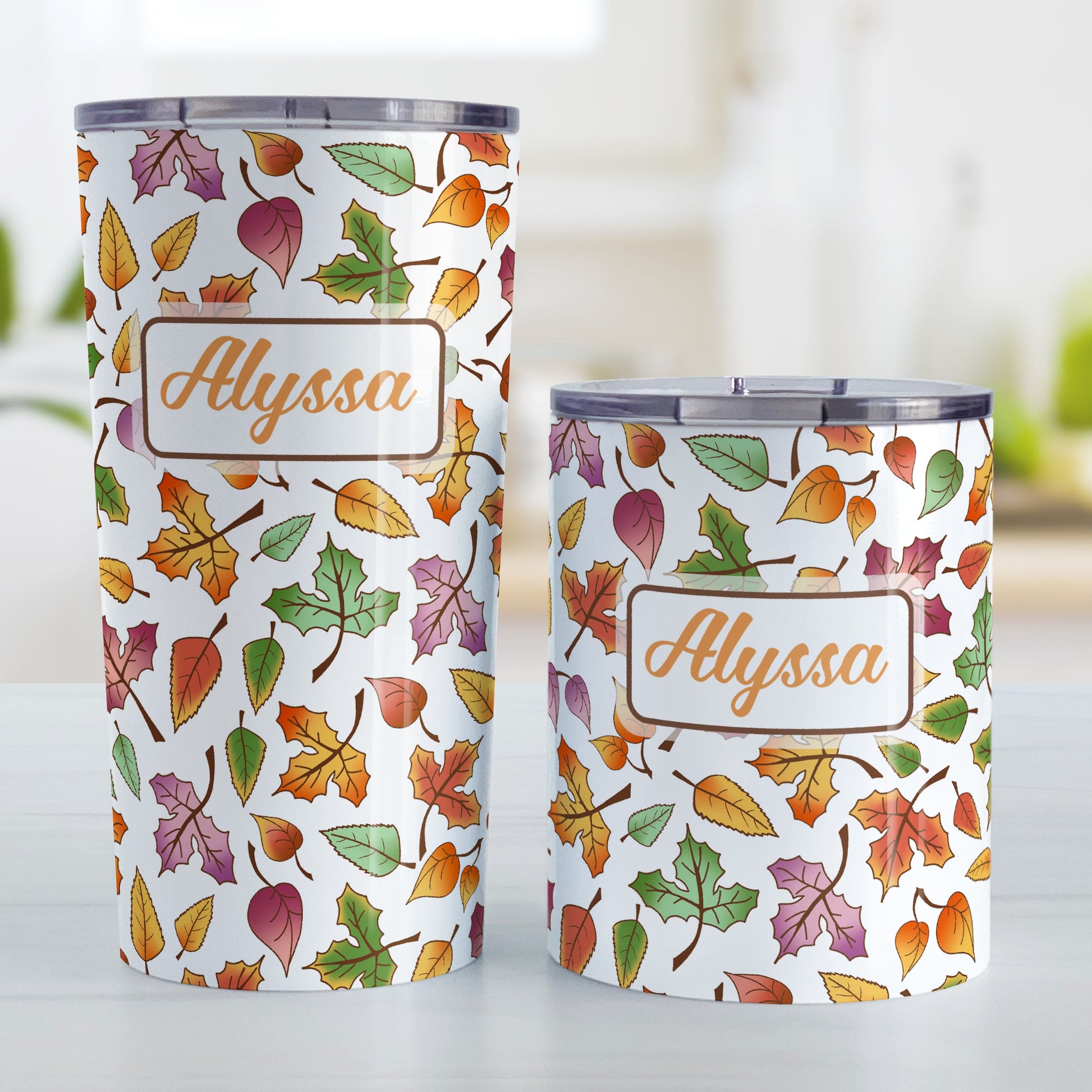 Personalized Changing Leaves Fall Tumbler Cups (20oz or 10oz) at Amy's Coffee Mugs. Stainless steel insulated tumbler cups designed with a fall themed pattern of leaves changing colors, as they do at the beginning of autumn, that wraps around the cups. Your personalized name is custom printed in an orange script font in a white rectangle over the leaves pattern. Photo shows both sized cups next to each other.