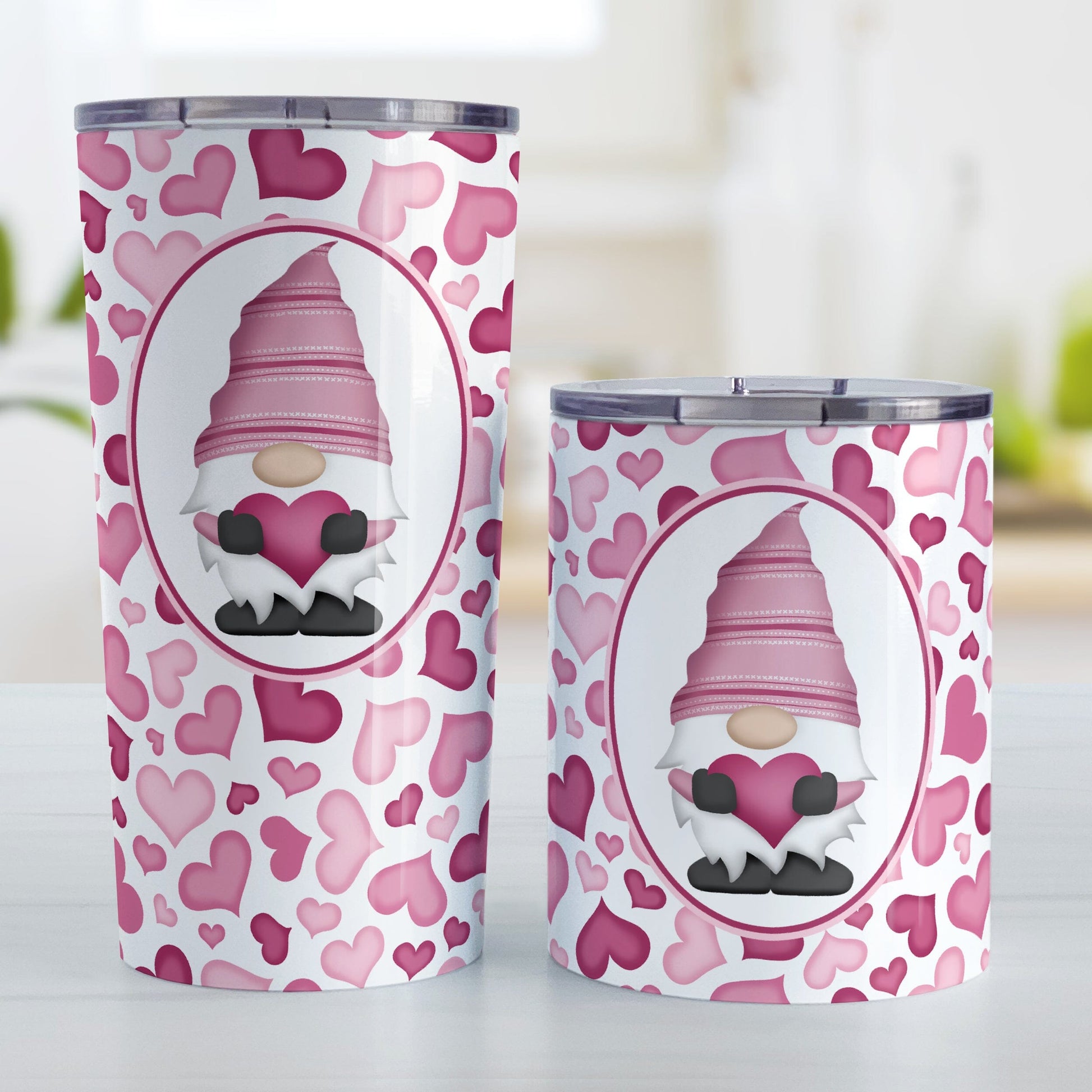 Pink Gnome Hearts Tumbler Cup (20oz or 10oz) at Amy's Coffee Mugs. Stainless steel tumbler cups designed with an adorable pink gnome holding a heart in a white oval over a pattern of cute hearts in different shades of pink that wrap around the cups. Photo shows both sized cups next to each other on a table.