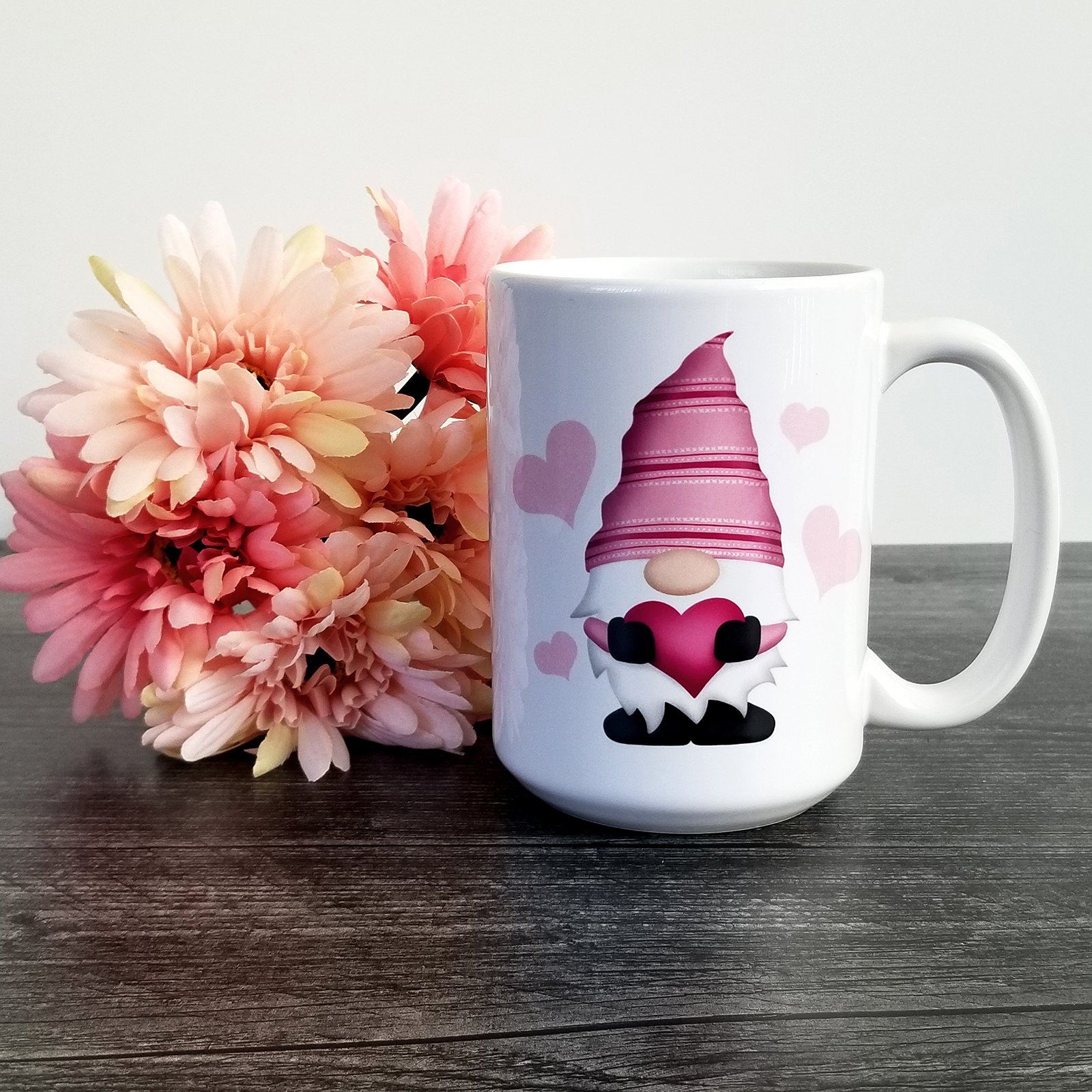 Pink Heart Gnome Mug (15oz) on a weathered wood table next to flowers. Pink Heart Gnome Mug available at Amy's Coffee Mugs