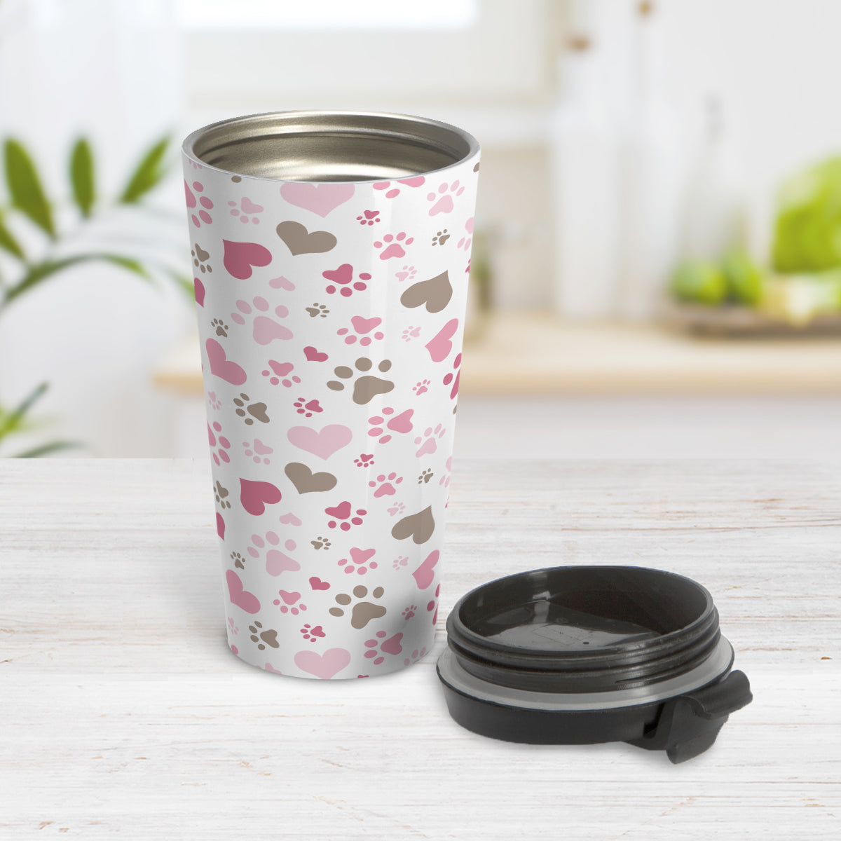 Pink and Hearts Paw Prints Travel Mug (15oz) at Amy's Coffee Mugs. A stainless steel travel mug designed with a pattern of hearts and paw prints in brown and different shades of pink that wraps around the mug. This travel mug is perfect for people love dogs and cute paw print designs. Photo shows the cup with the lid on the table beside it.